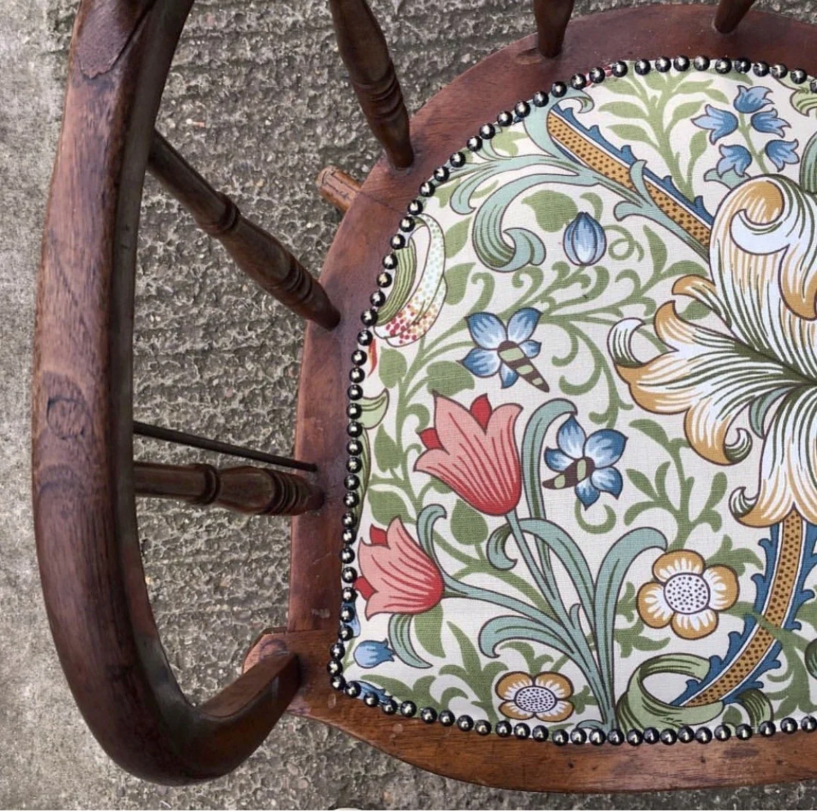 Close-up of a wooden chair with a floral upholstered seat featuring pink, blue, orange, and green colors, with nailhead trim around the edge.