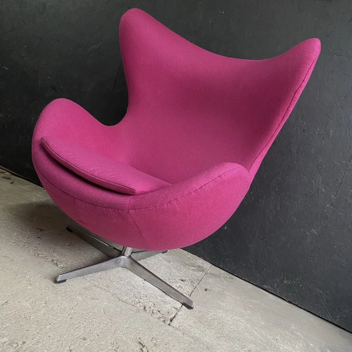 Pink mid-century modern style lounge chair with a high back and curved armrests, set against a black wall and concrete floor.