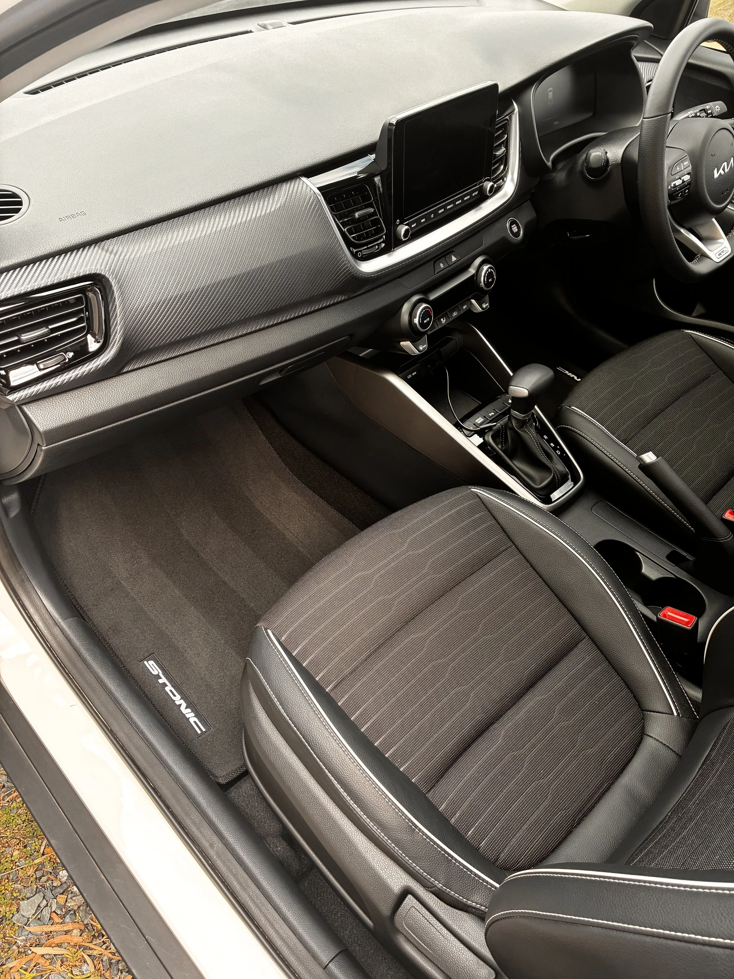 Inside a modern car showing the front passenger seat, dashboard, steering wheel, touchscreen display, air vents, and floor mat with the word 'STONIC' on it.