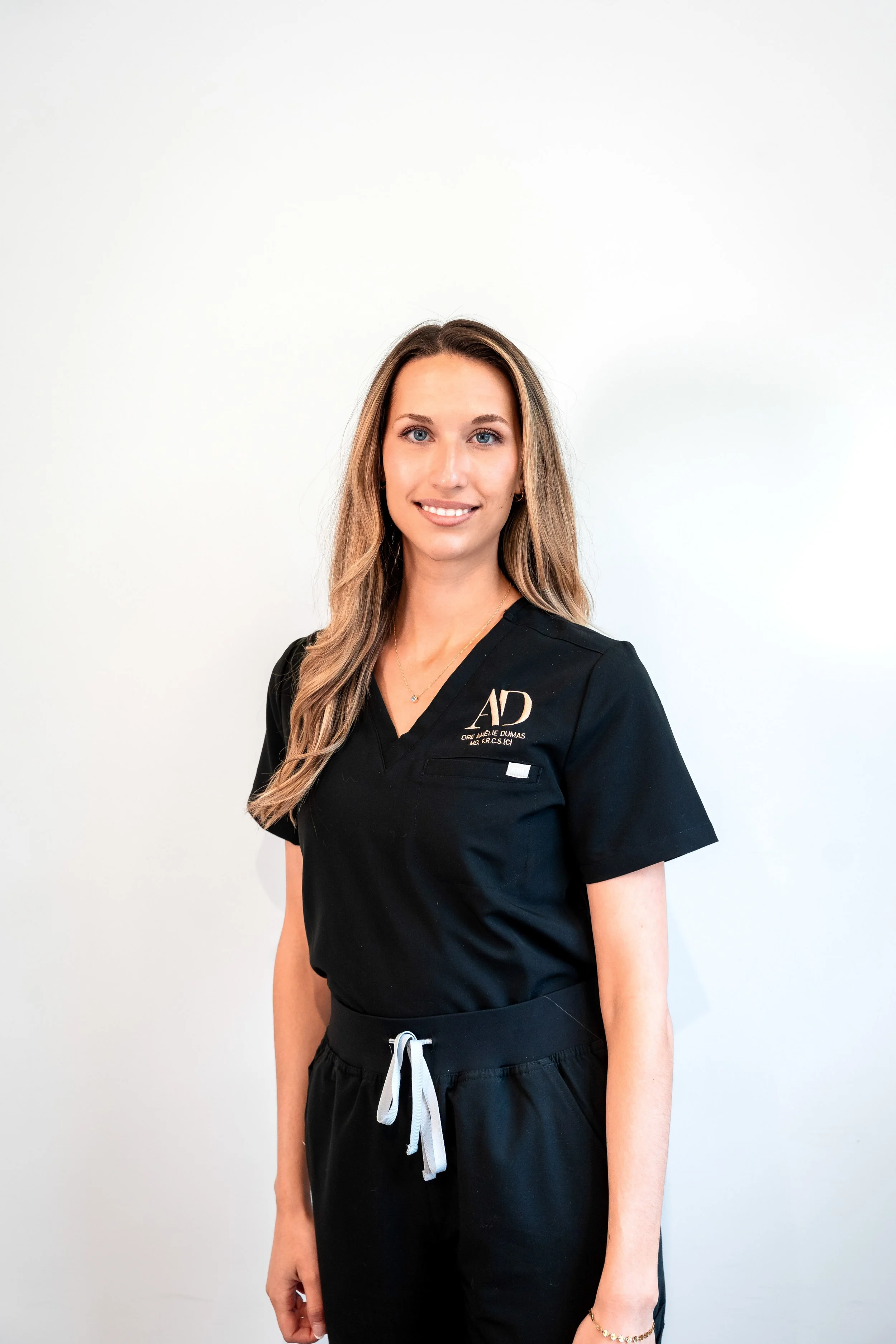 Une femme porte un uniforme noir avec des initiales 'AD' brodées, sourit et pose contre un fond blanc.