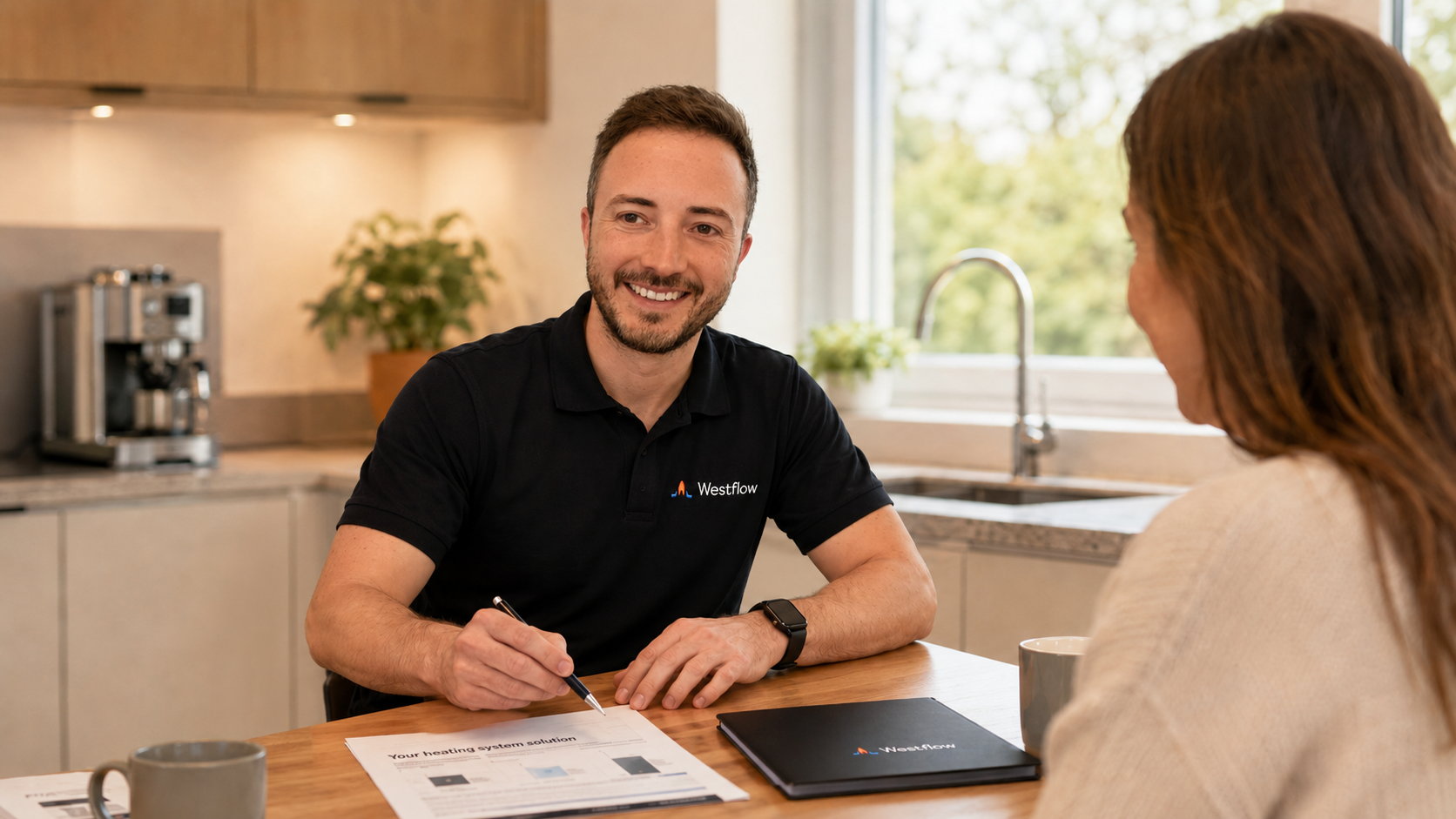 A man and woman having a discussion at a kitchen table. The man is smiling and holding a pen, wearing a black polo shirt with 'Westflow' logo. Papers and a laptop are on the table.