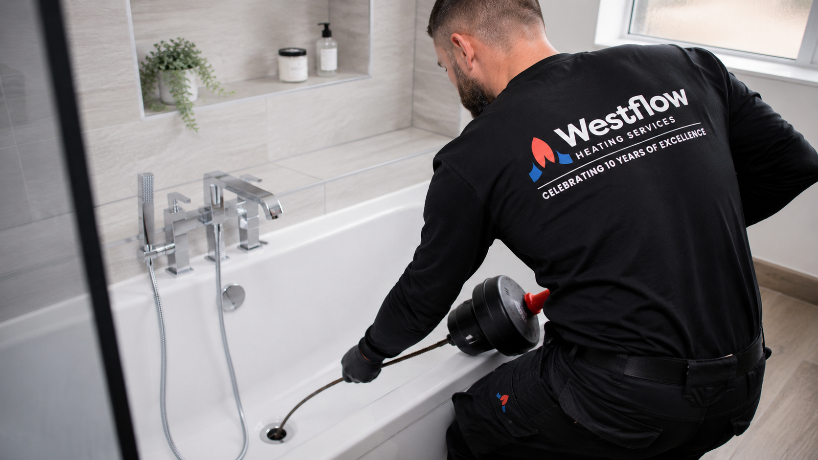 A plumber using a drain auger to clear a bathtub drain in a bathroom with beige wall tiles and a window.