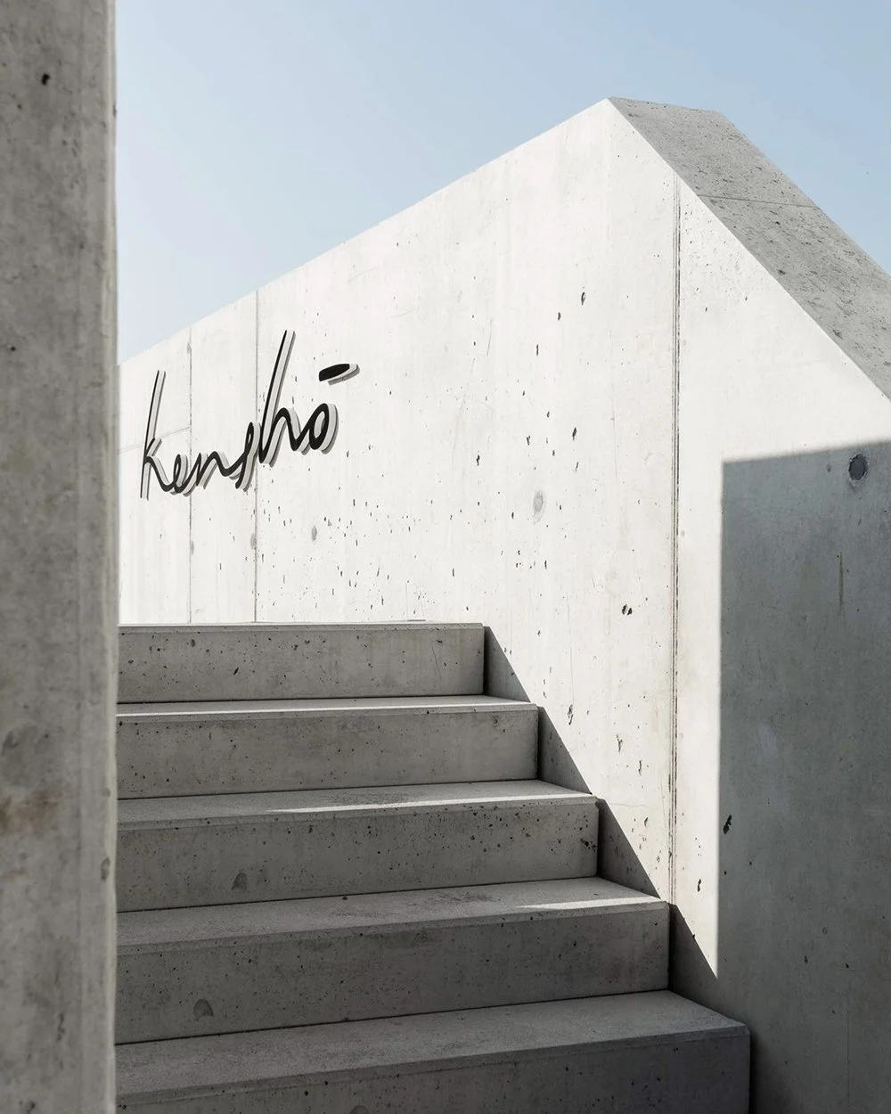 Foto von einer modernen, minimalistischen Treppe aus Beton, die zu einer weißen Betonwand mit dem Wort 'kerhä' in geschwungener Schrift führt.