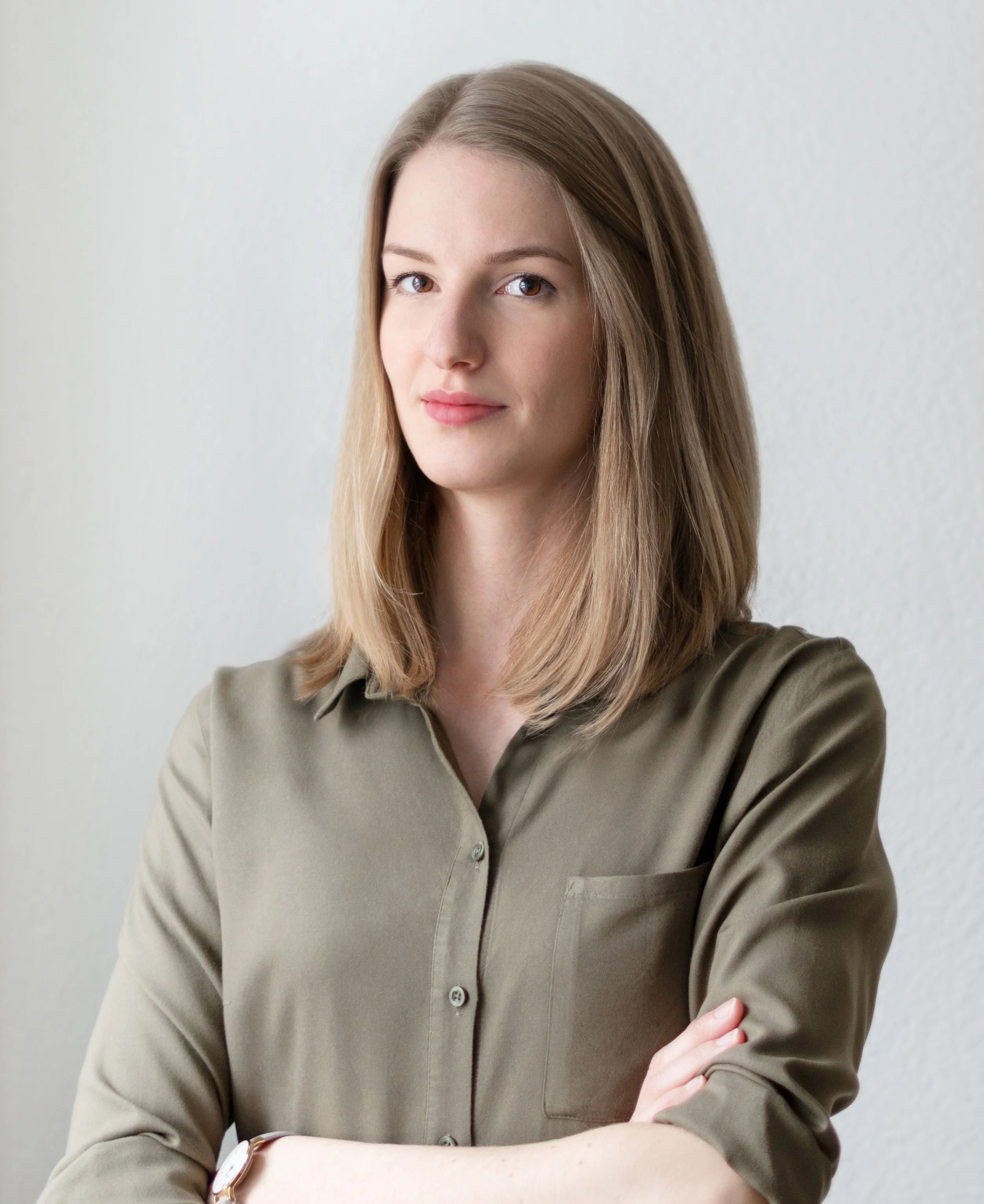 Nahaufnahme einer jungen Frau mit braunen Haaren, die ein khakifarbenes Hemd trägt und die Arme verschränkt hat, vor einer weißen Wand.
