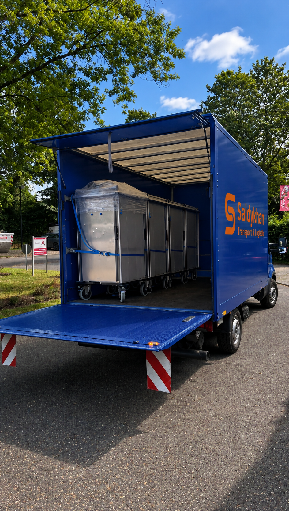 Ein Mann in einer gelben Jacke lädt Pakete in einen Lieferwagen. Es ist Abendzeit, der Himmel ist bewölkt, und die Straße ist nass.