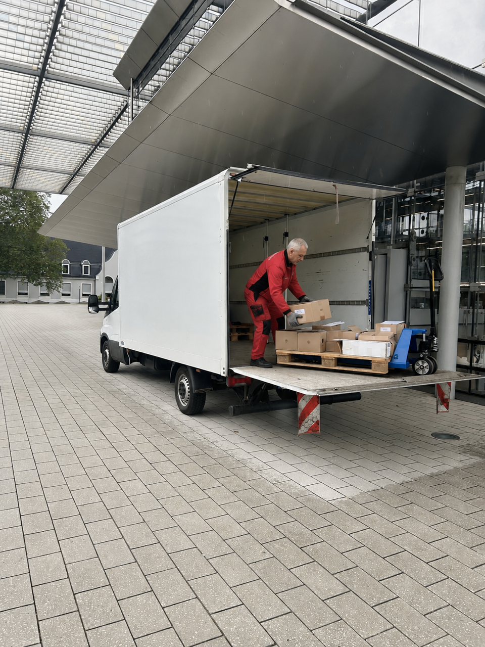 Ein Mann in einer gelben Jacke lädt Pakete in einen Lieferwagen. Es ist Abendzeit, der Himmel ist bewölkt, und die Straße ist nass.