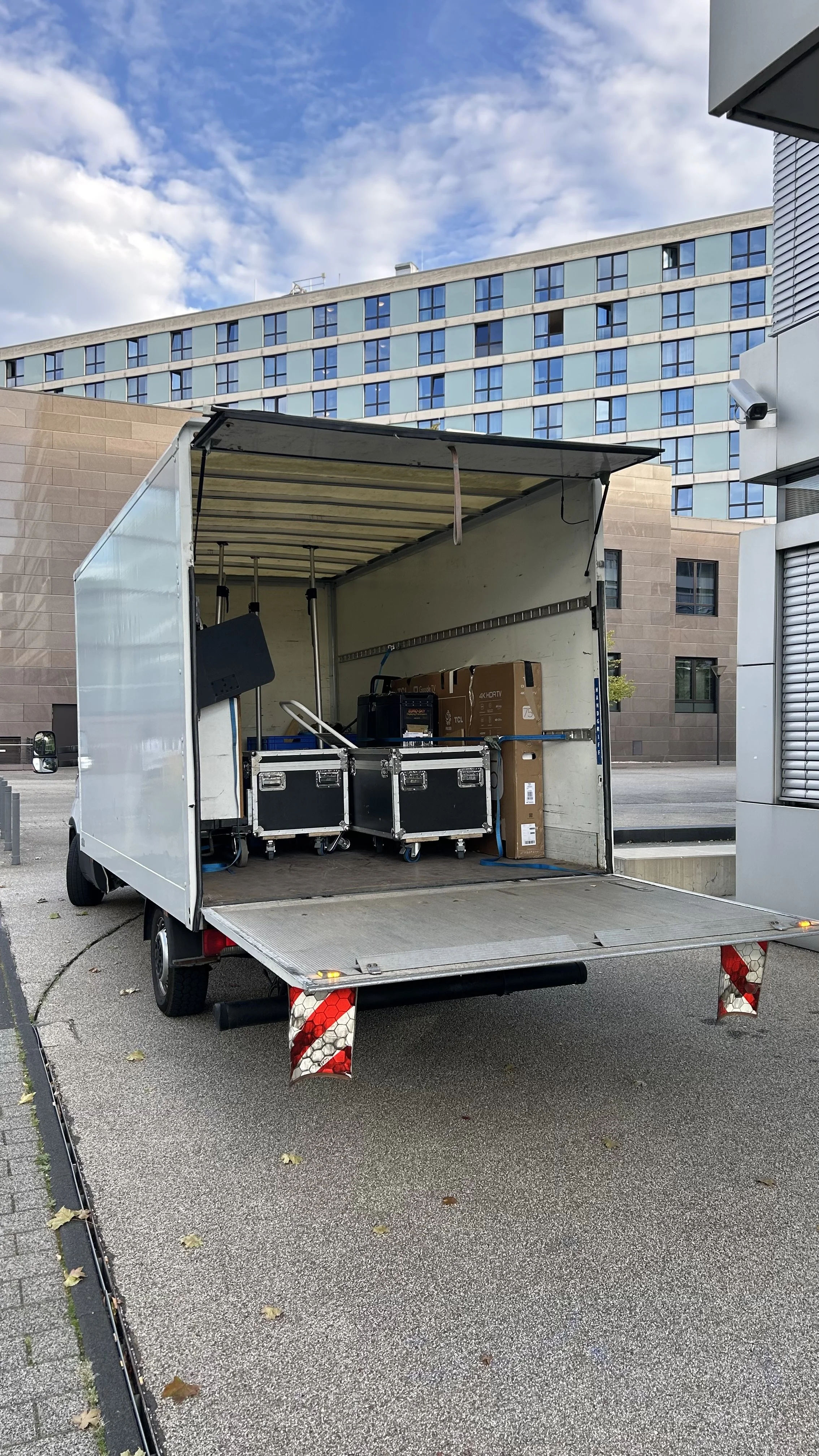 Ein offener Güterwagen mit Kartons, Kisten und Verpackungsmaterialien, bereit zum Transport, auf einer Stadtrandstraße mit modernen Gebäuden im Hintergrund.