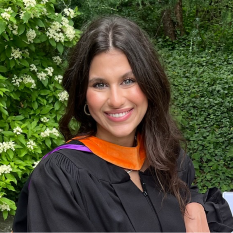 Woman in graduation gown outdoors, smiling.