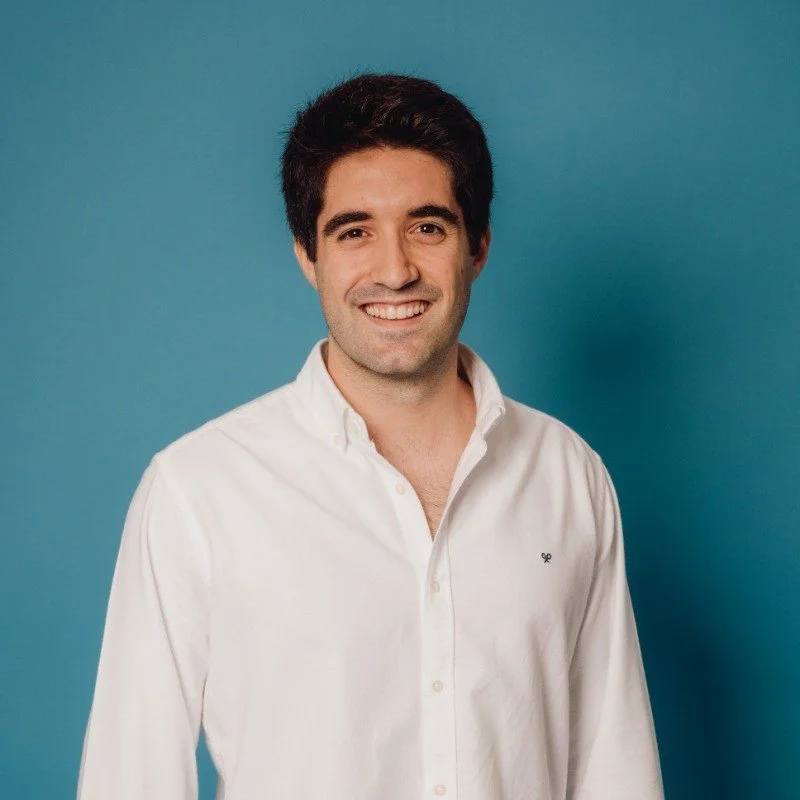 Portrait of a young man with dark hair, smiling, wearing a white button-up shirt, standing against a blue background.