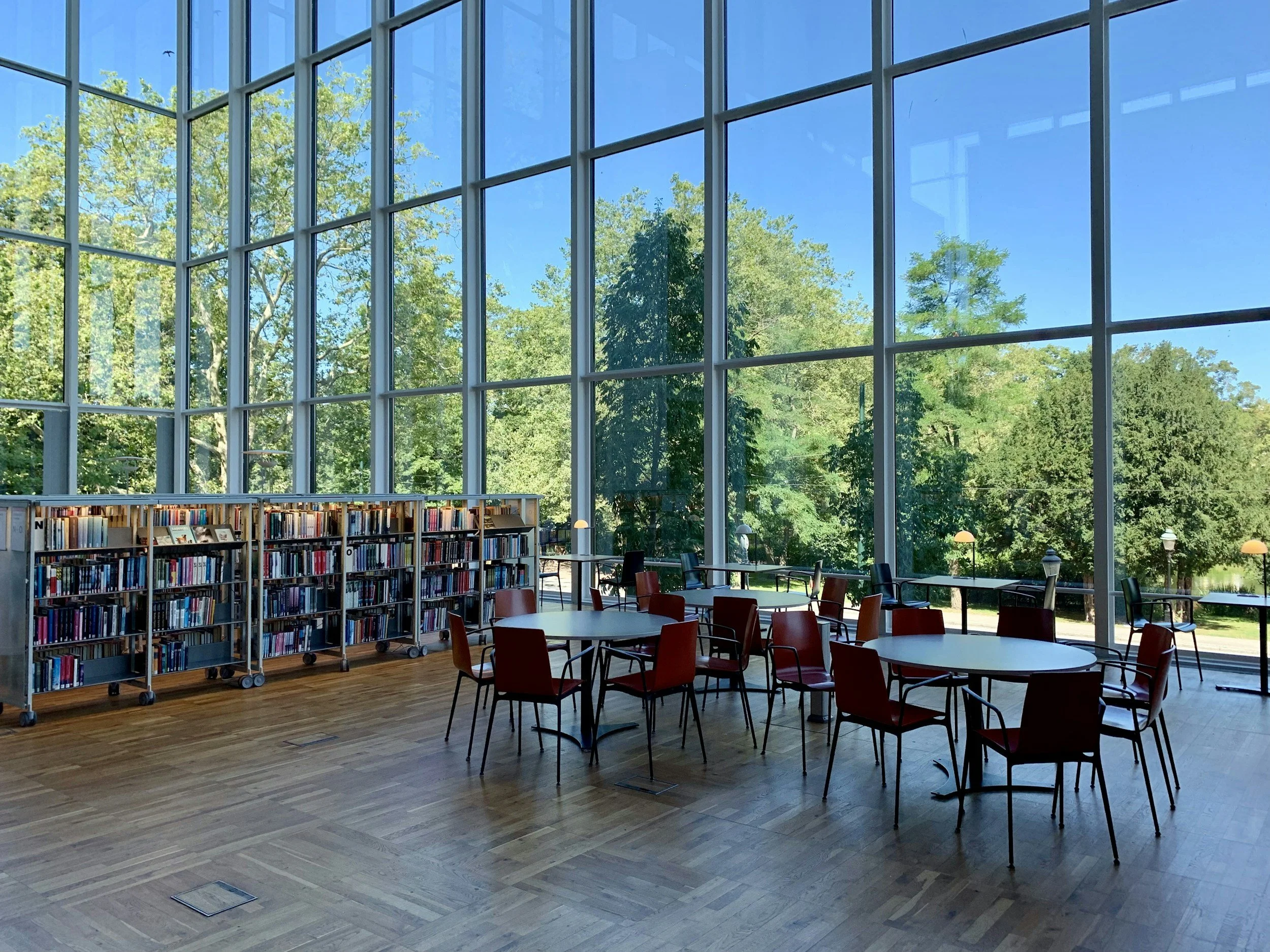 Interior of a modern library or reading room with large glass windows showing green trees outside. There are bookshelves on wheels, tables, and chairs inside.