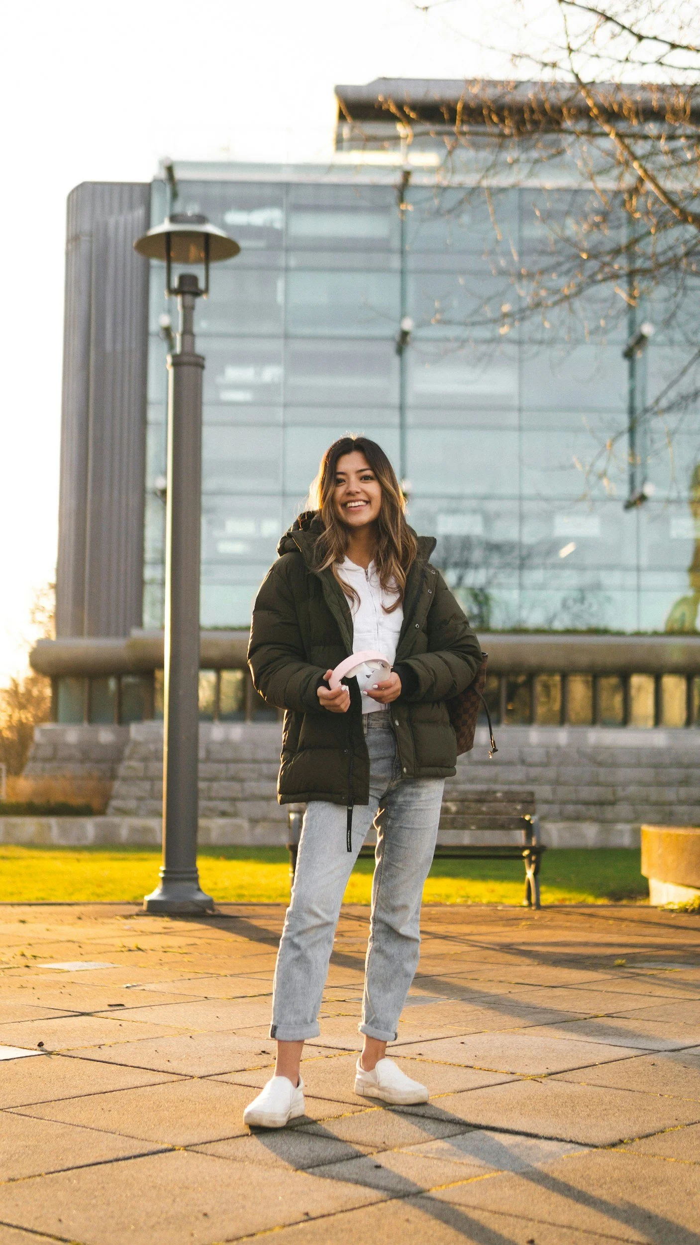 A young woman standing outdoors on a sidewalk during sunset, holding headphones, wearing a jacket, white shirt, gray jeans, and white sneakers, with modern glass building and trees in the background.