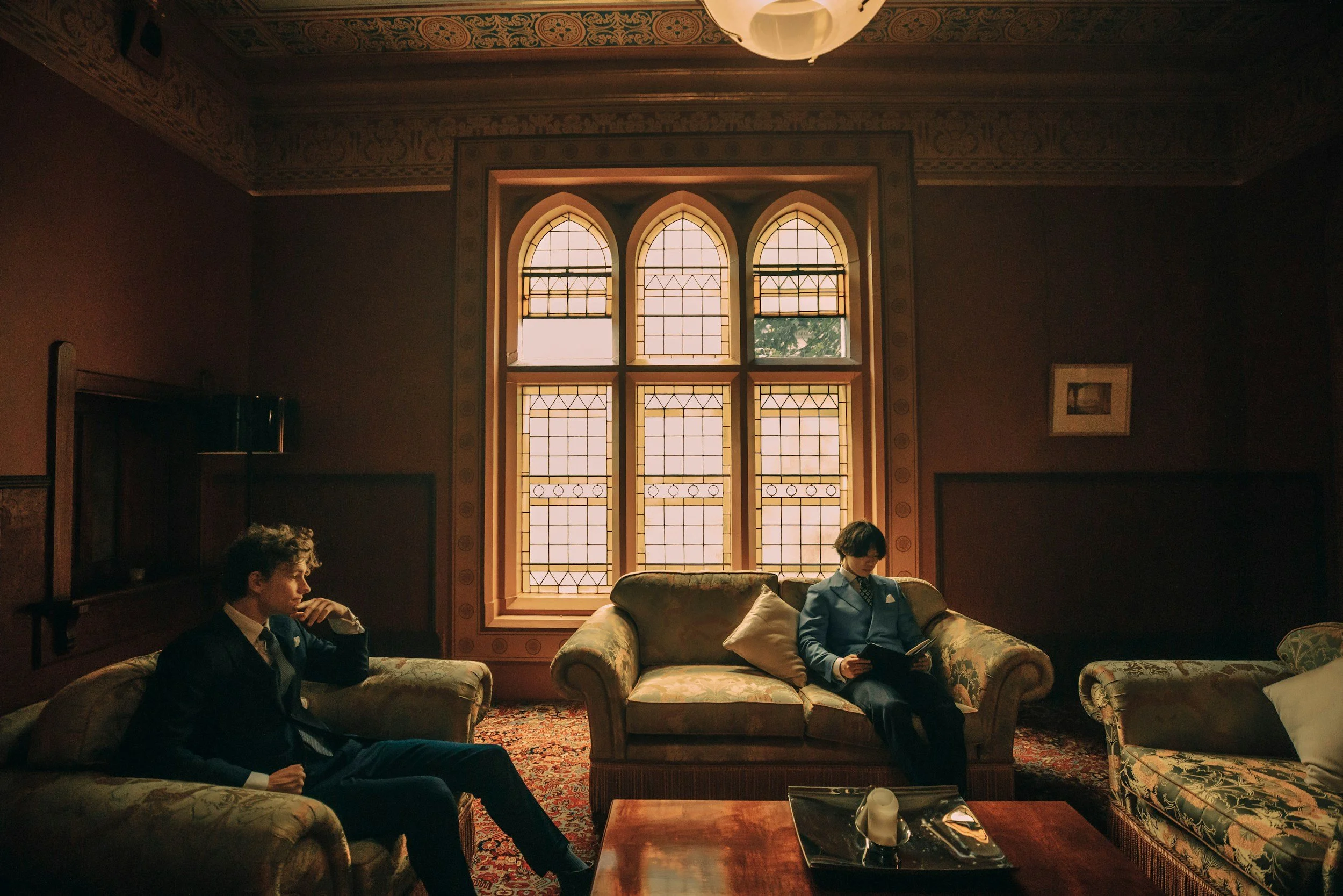 Two men dressed in suits sitting on floral patterned sofas in a dimly lit room with a large stained glass window and ornate ceiling trim, one reading a book and the other resting with his hand on his face.