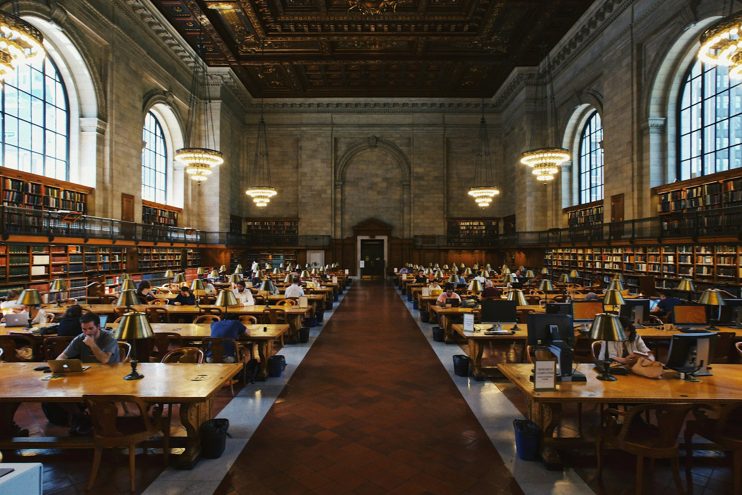 A large library reading room with rows of wooden tables and green-shaded lamps, high arched windows, high ceiling with intricate gold and dark wood details, and multiple chandeliers with hanging lights. People are seated at the tables working on laptops and reading.