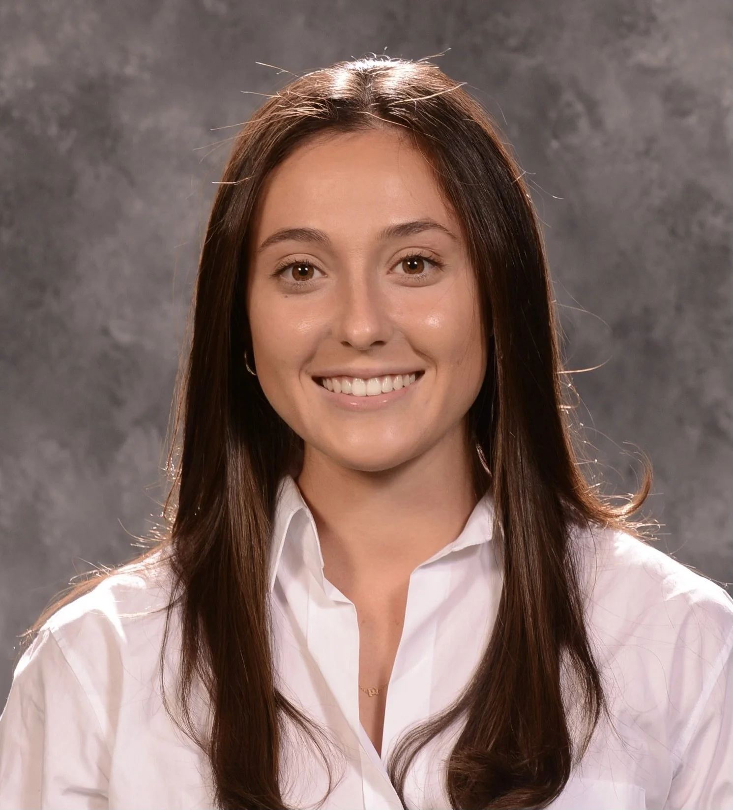 Young woman with long brown hair, wearing a white shirt, smiling, posing against a gray textured background.