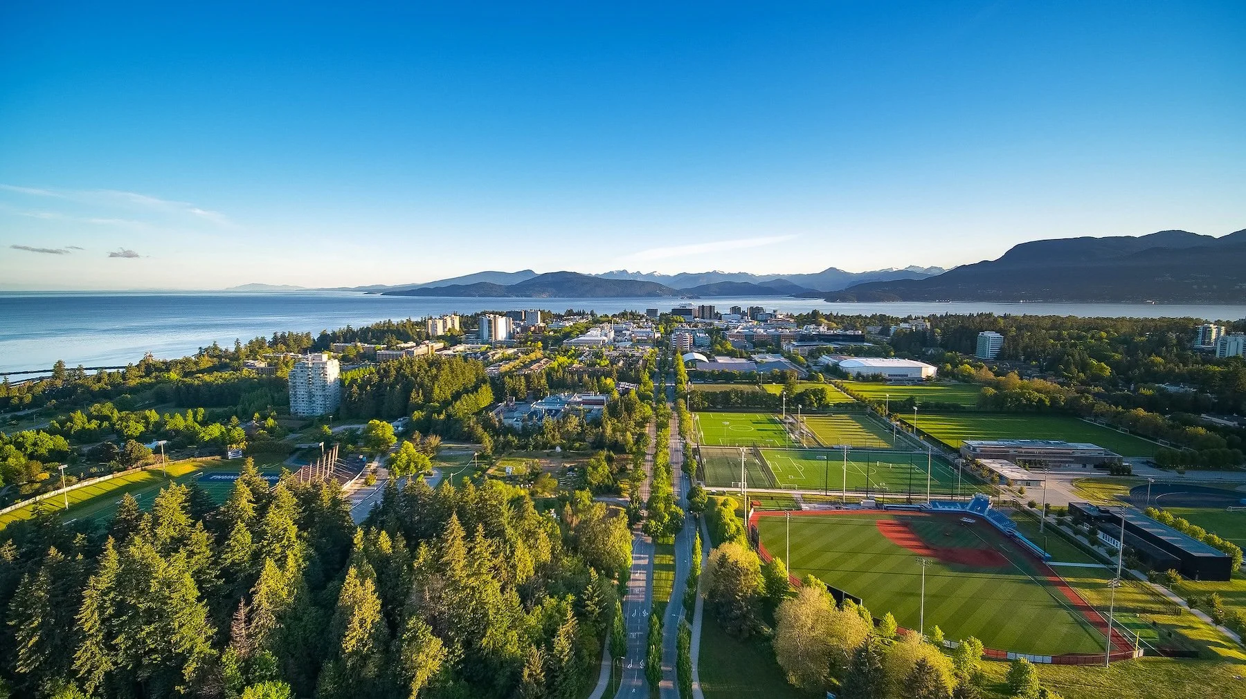 Aerial view of a city with parks, sports fields, buildings, and a large body of water with mountains in the background under a clear blue sky.