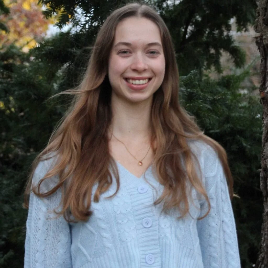 A young woman with long, wavy brown hair and fair skin is smiling outdoors. She is wearing a light blue cardigan with large buttons and a small gold necklace, with greenery and trees in the background.