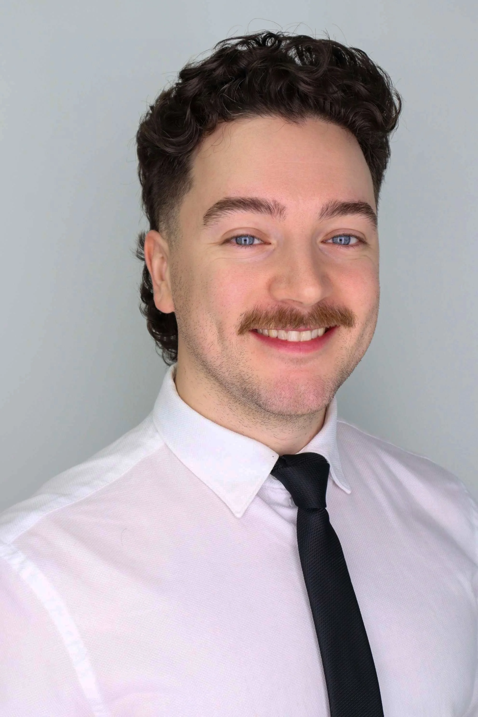 Close-up of a young man with dark curly hair, blue eyes, and a mustache, wearing a white dress shirt and black tie, smiling against a plain light gray background.