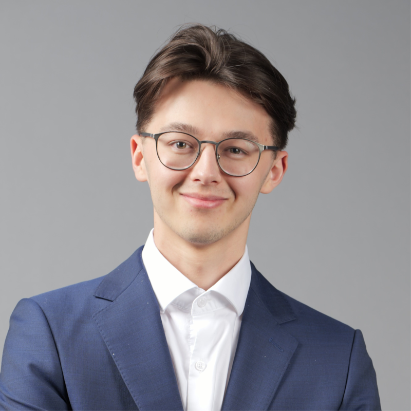 Portrait of a young man with light skin, brown hair, glasses, wearing a navy blue suit and white shirt, smiling against a gray background.
