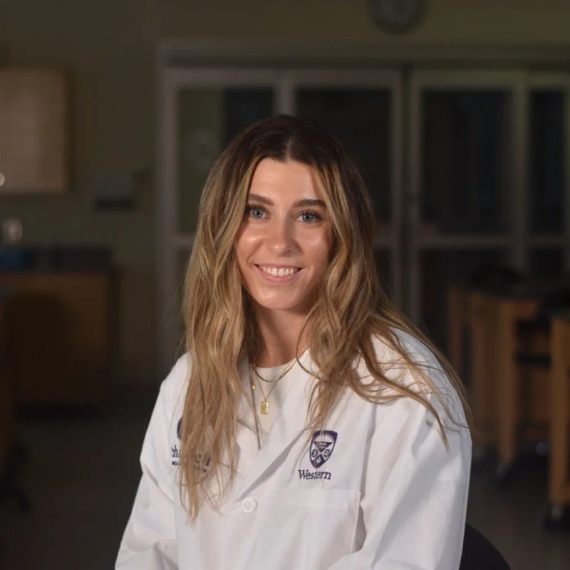 A young woman with long, wavy, blonde hair smiling, wearing a white lab coat with a logo and text that says 'Western', in a laboratory or classroom setting.