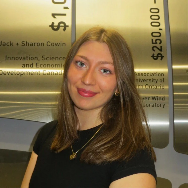 A young woman with long light brown hair, wearing a black top and gold jewelry, smiling at the camera, with a metallic plaque in the background showing award information.