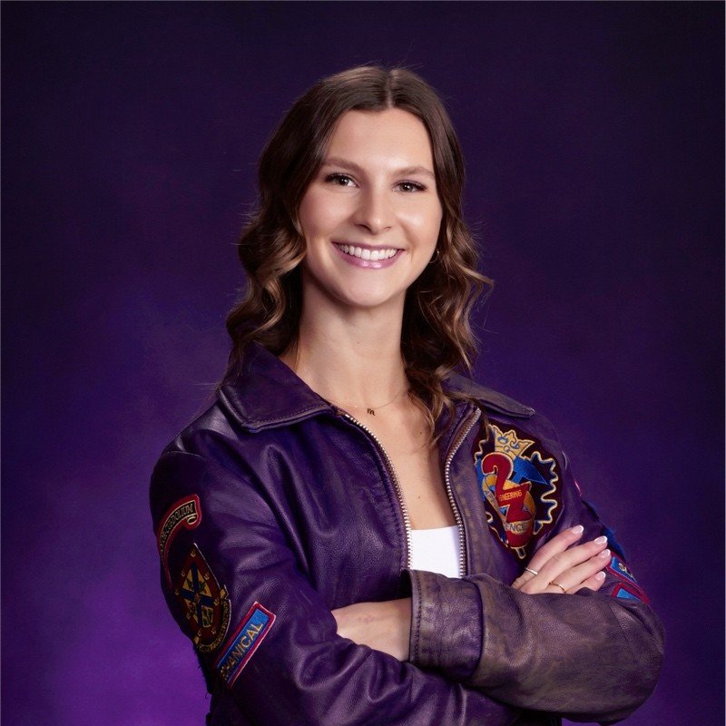 A woman with brown hair styled in loose waves, smiling with teeth showing, wearing a purple NASA jacket with patches, standing with arms crossed against a purple background.