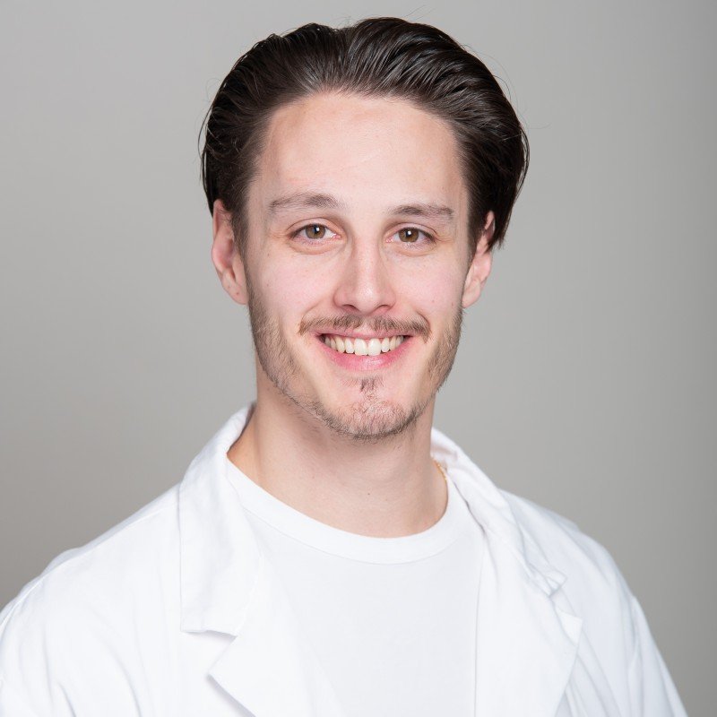 A young man with brown hair, styled back, wearing a white shirt, smiling at the camera, with a neutral gray background.