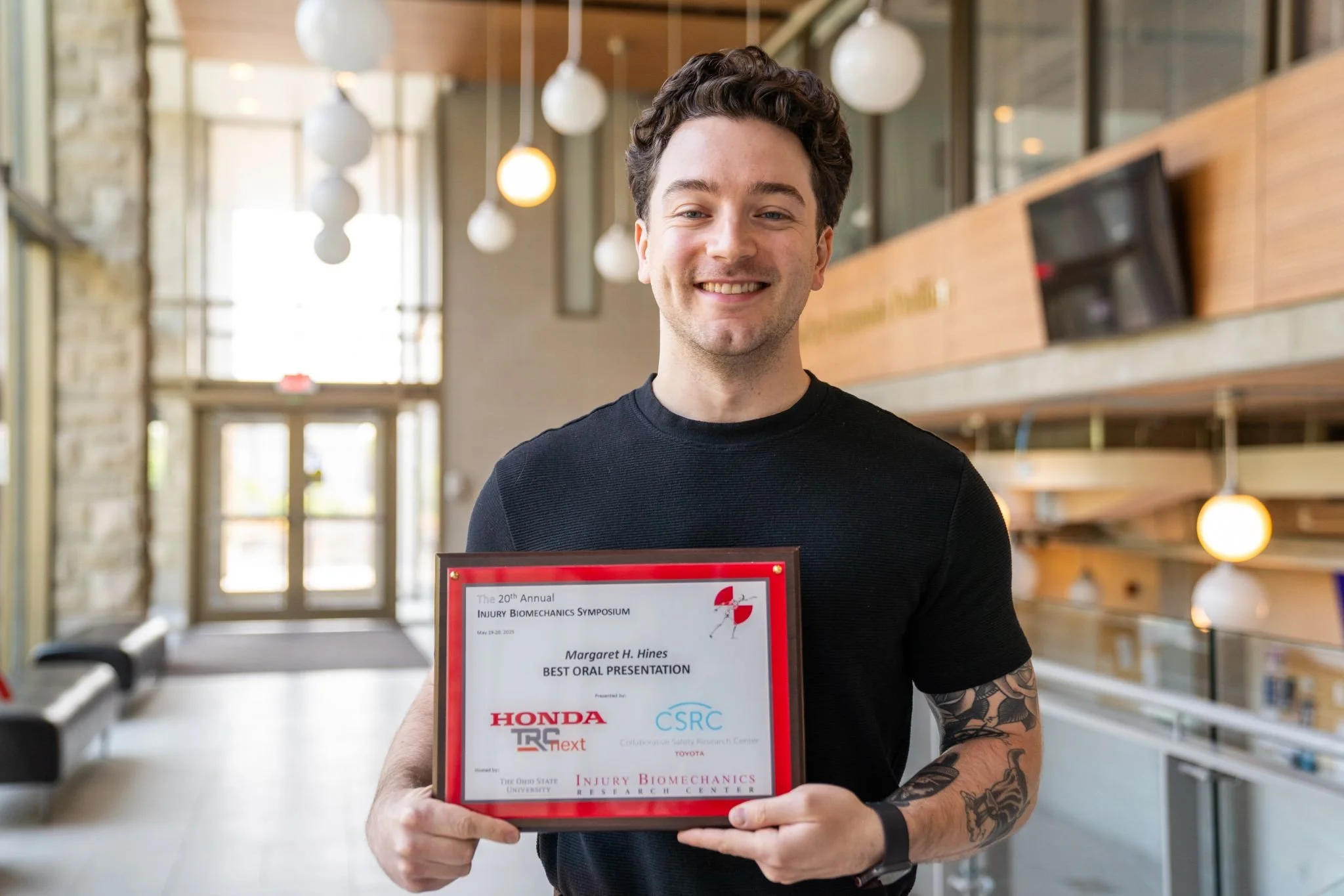 Man smiling while holding an award certificate at a conference or seminar in a modern indoor space with hanging lights and large windows.