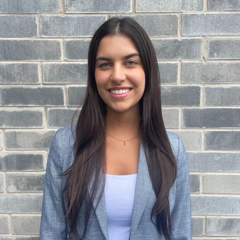 A young woman with long dark hair and a bright smile, wearing a grey blazer over a white top, standing in front of a brick wall.