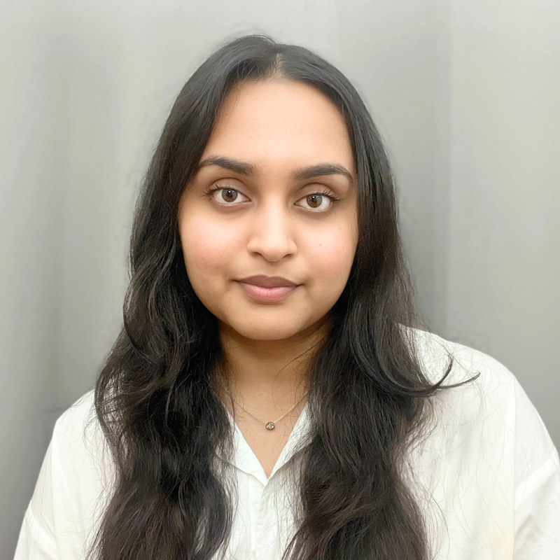 Portrait of a young woman with long dark hair, wearing a white top and a delicate necklace, standing in front of a plain light gray background.