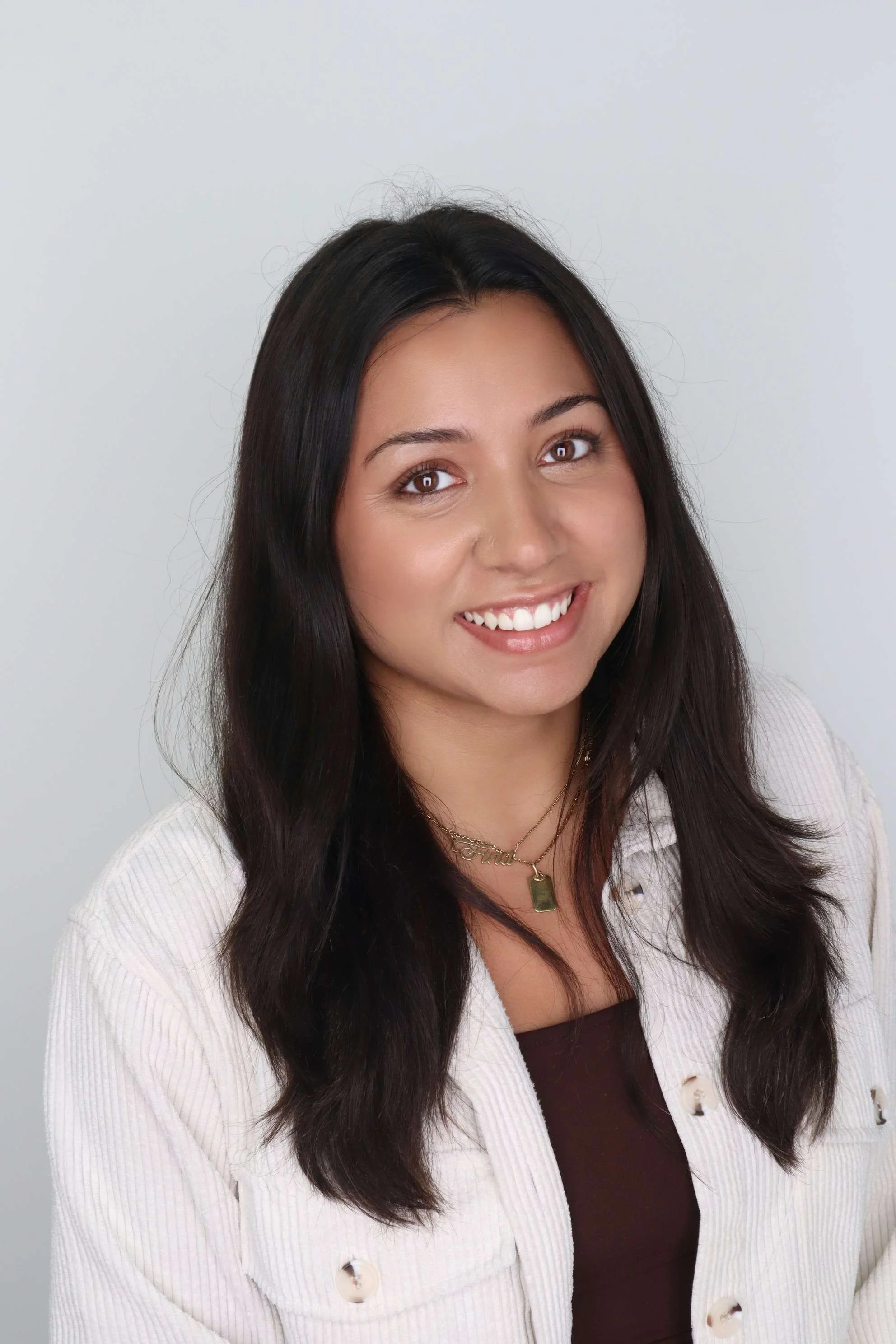 A smiling woman with long dark hair, wearing a white blazer over a dark top, accessorized with a gold necklace with a pendant.