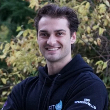 A young man with dark hair smiling in front of green and yellow foliage, wearing a navy blue hoodie.