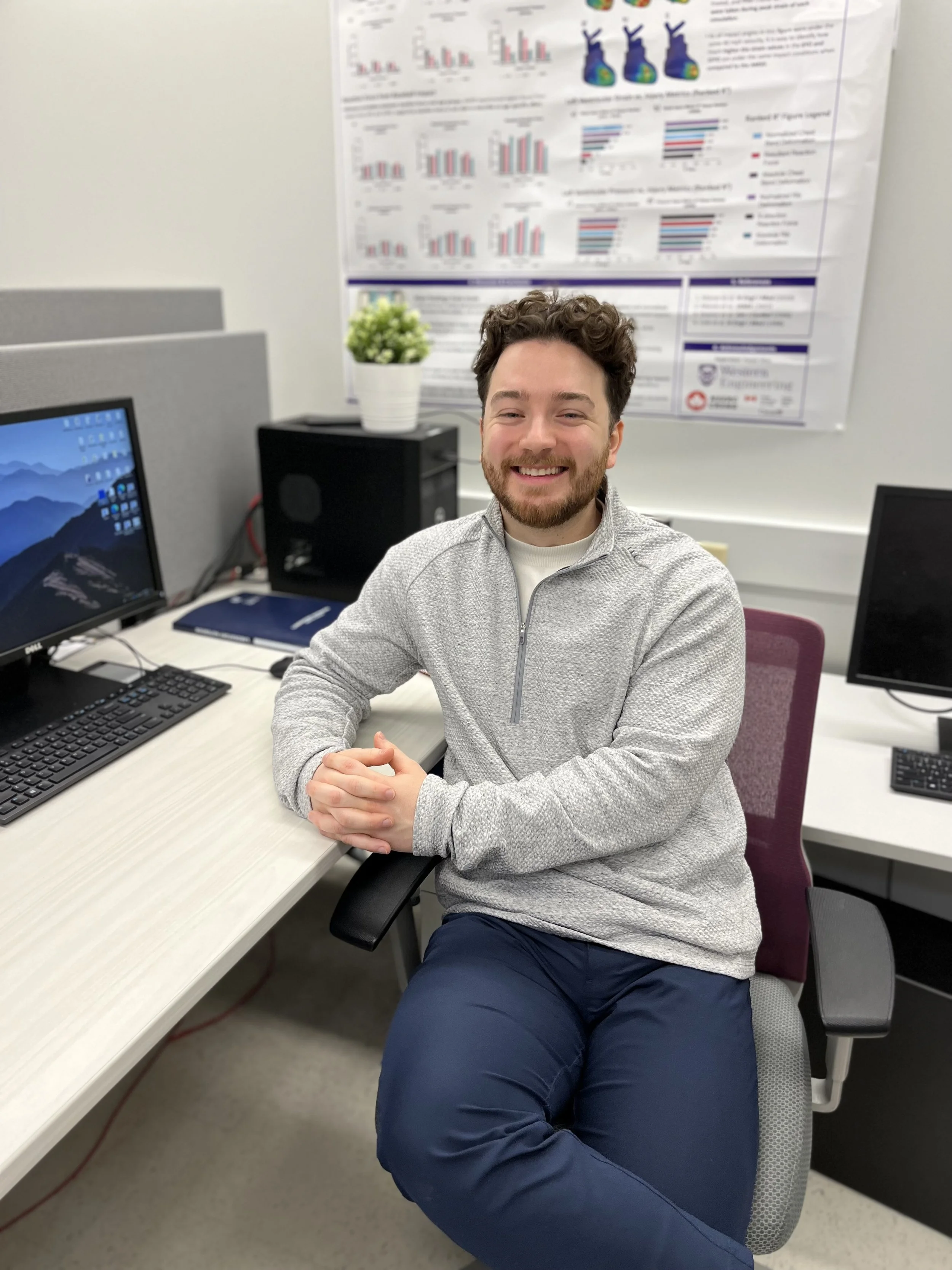 A man with curly hair and a beard smiling at the camera, sitting at a desk in an office. He is wearing a light gray zip-up sweater and navy blue pants. On the desk are two computer monitors, a black keyboard, and a small blue notebook. Behind him, there is a large poster with charts and graphs on the wall, a potted plant, and a computer tower.
