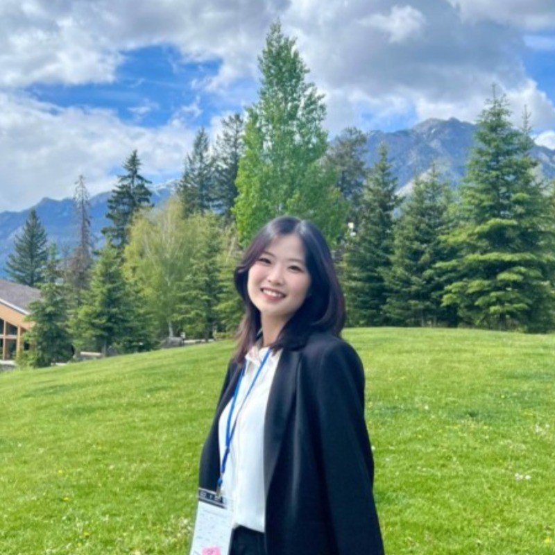 Smiling young woman with dark hair wearing a white shirt, black blazer, and conference badge, standing on a green grassy area with trees and mountains in the background under a partly cloudy sky.
