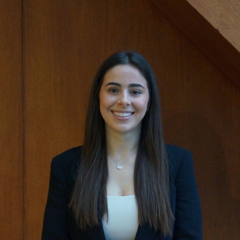 A young woman with long dark hair smiling, wearing a black blazer and a white top, standing in front of a wooden background.