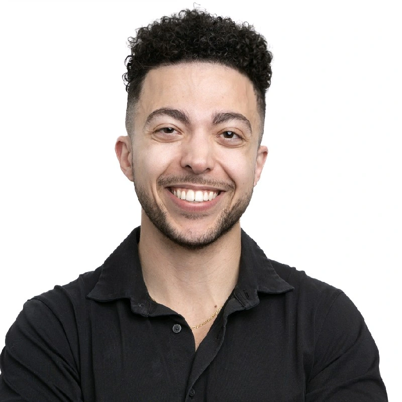 A young man with curly dark hair, a light beard, and a big smile, wearing a black shirt and a gold chain, standing against a white background.