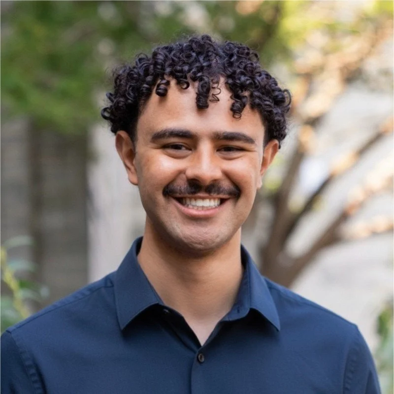 Young man with curly dark hair and a mustache smiling outdoors, wearing a navy blue button-up shirt.