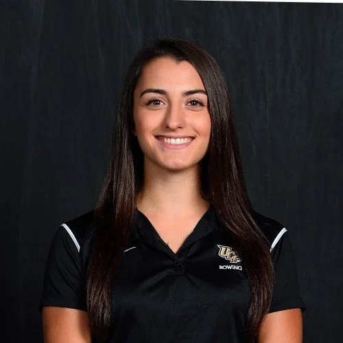 Young woman with long dark hair smiling, wearing a black sports shirt with a UCF rowing logo, against a dark background.