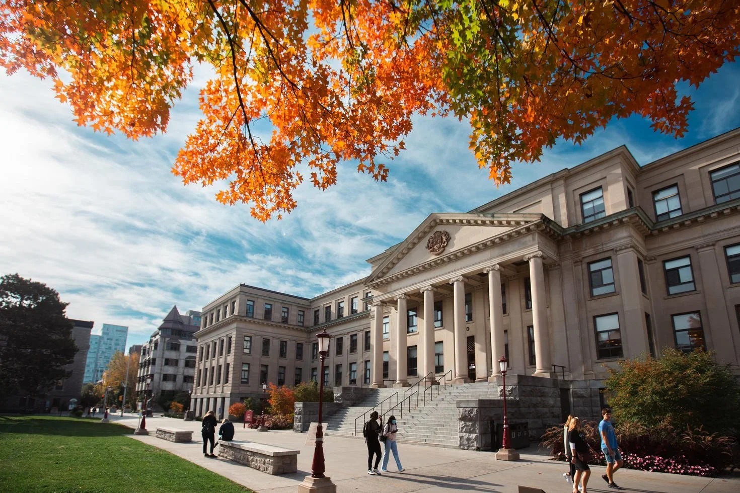 People walking on a university campus with a large historic building and trees with autumn leaves.