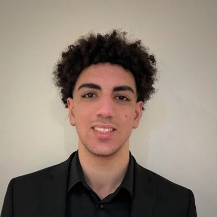 A young man with curly dark hair wearing a black suit and shirt, smiling slightly at the camera against a plain light-colored wall.