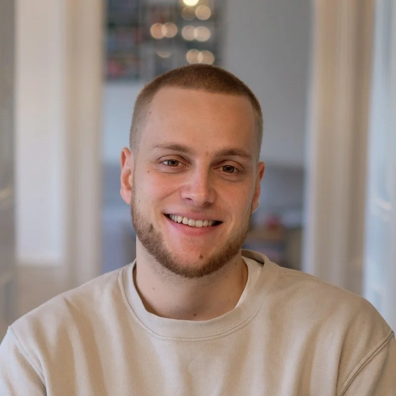 A young man with short blond hair, a light beard, and a beige sweatshirt smiling indoors with blurred background.