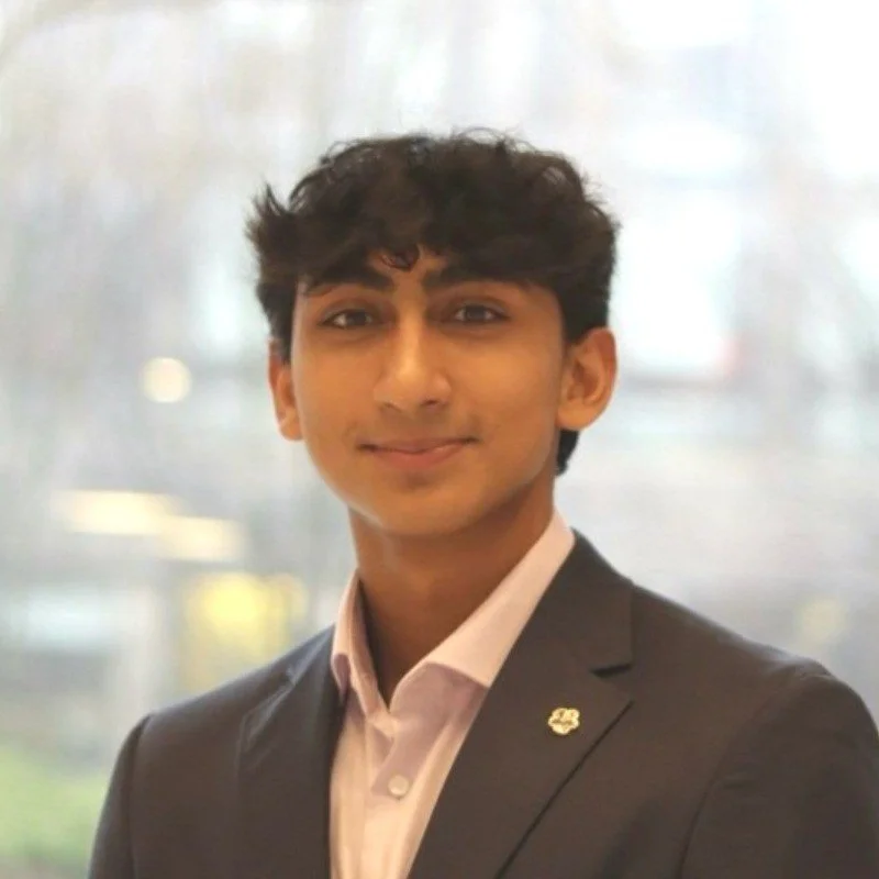 A young man in a suit and pink shirt, smiling, in front of a blurred indoor background.