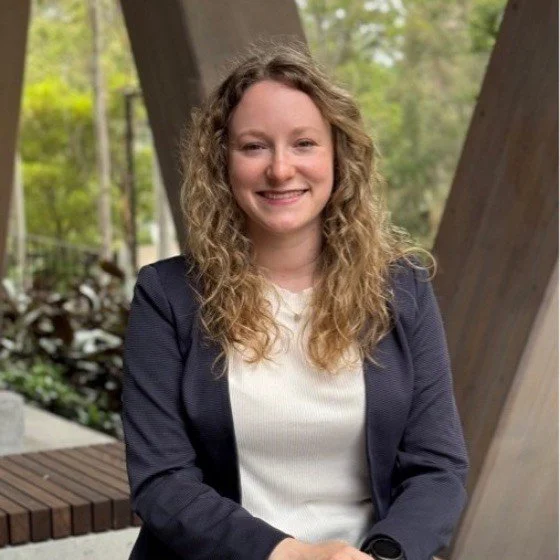 A woman with curly blonde hair and a big smile, wearing a navy blazer and white top, standing outdoors in front of a wooden structure and trees.
