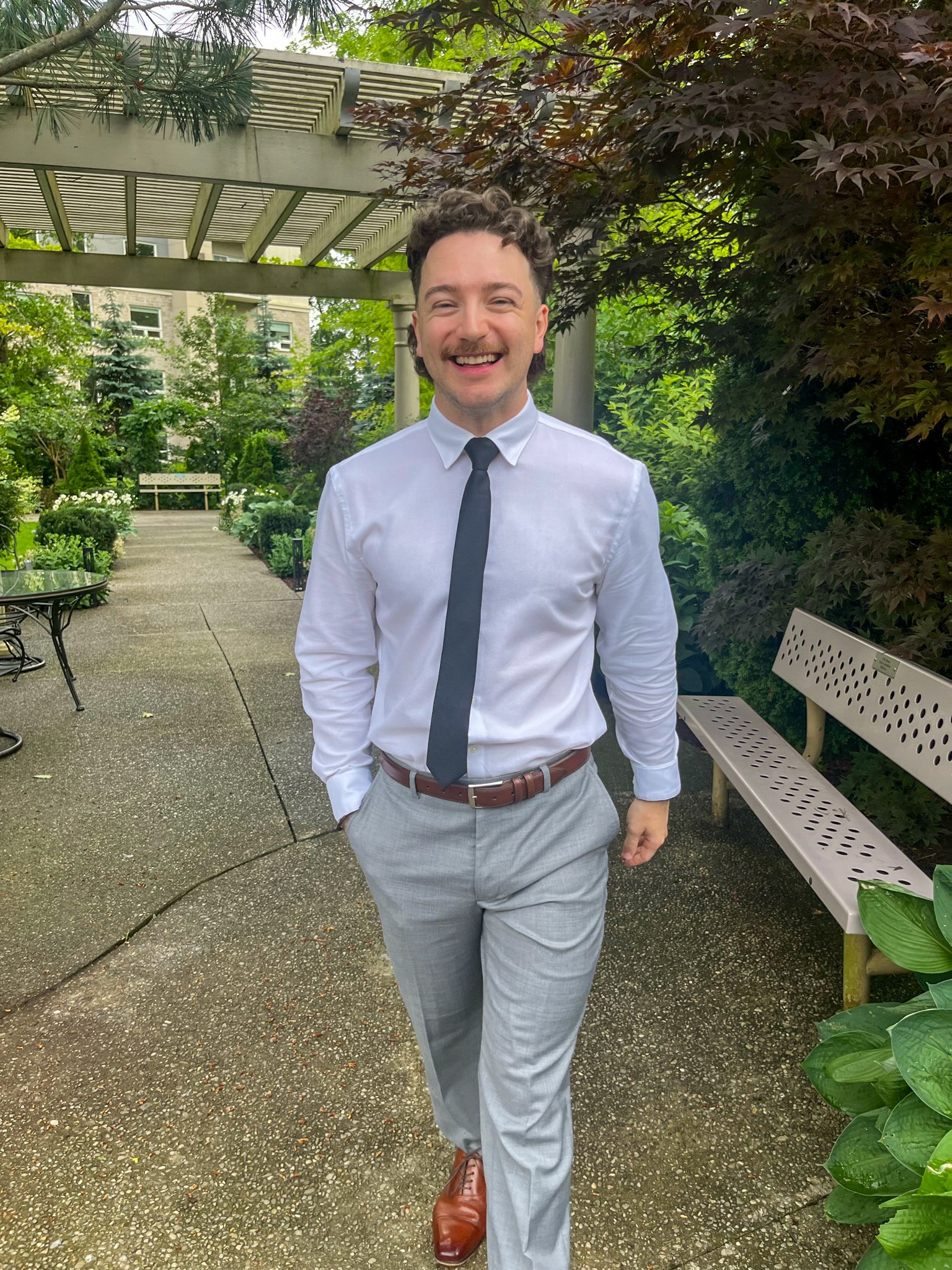 A man smiling and walking outdoors in a garden area with greenery, trees, plants, and park benches, wearing a white shirt, black tie, and light gray pants.