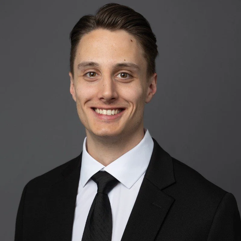 A young man with light skin, brown hair, and wearing a black suit, white shirt, and black tie, smiling against a gray background.