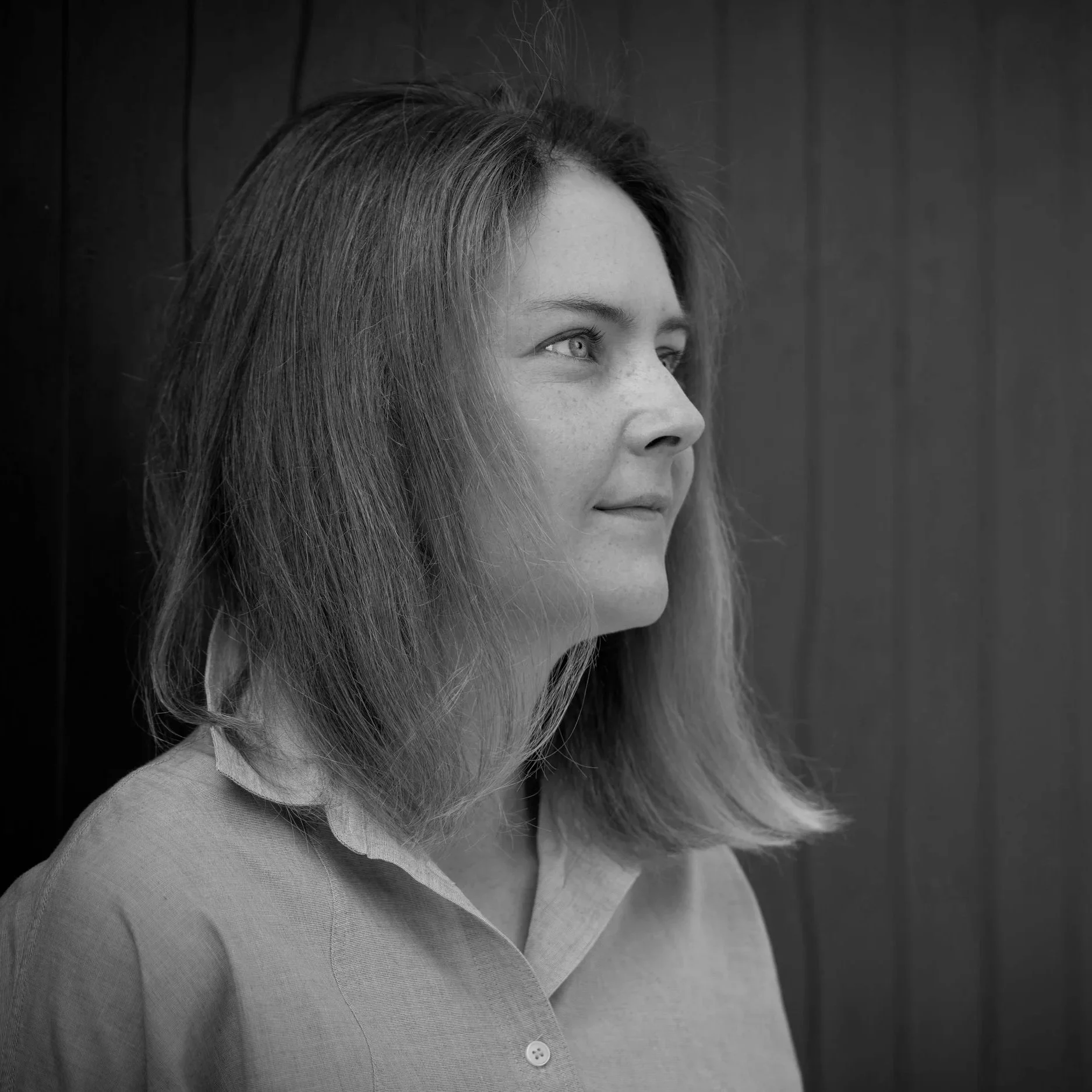 Close-up black-and-white photo of a woman with long hair, wearing a collared shirt, looking to the side against a wooden background.
