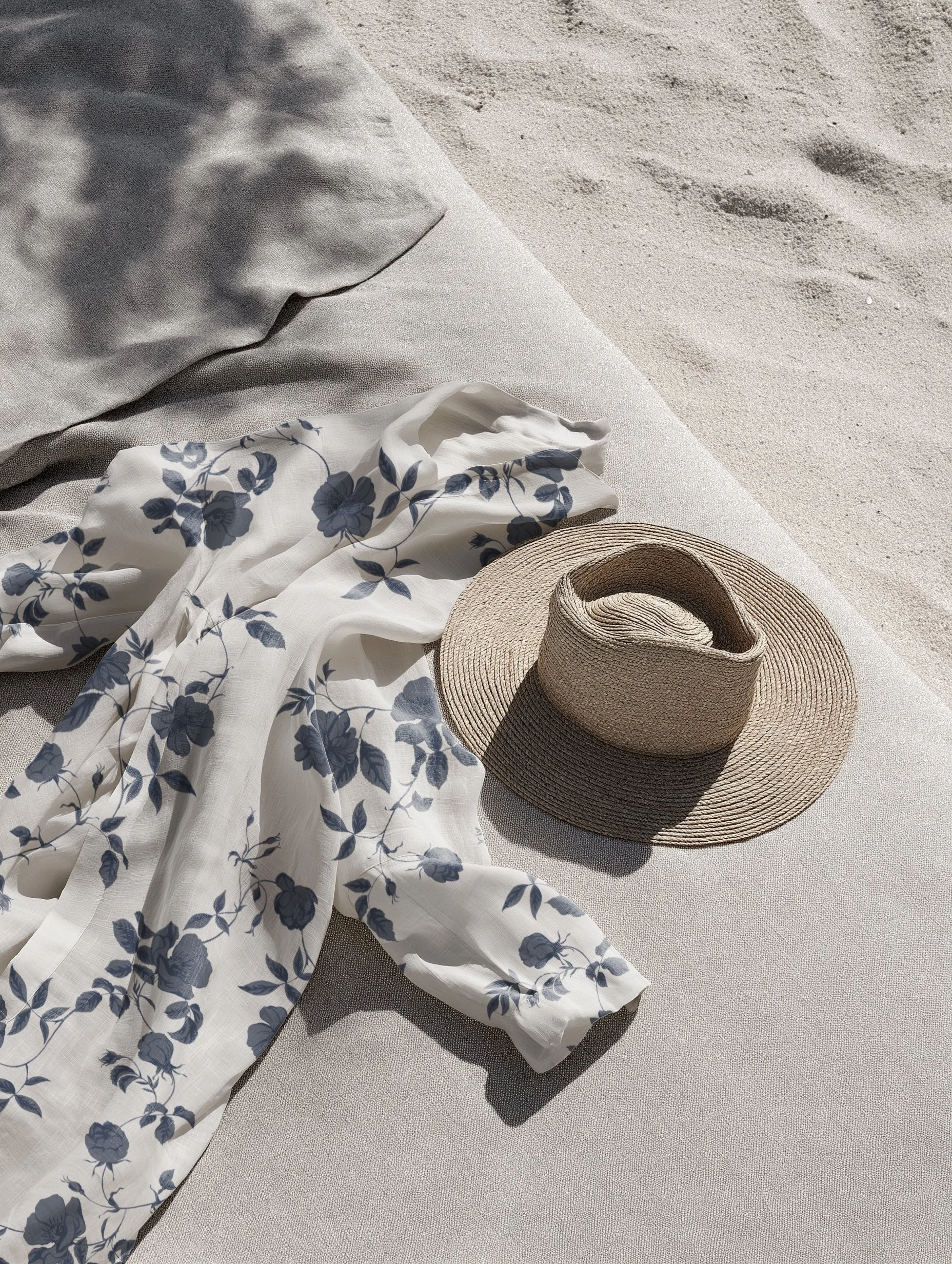 A beach scene with a beige towel, a white fabric with a blue floral pattern, a wide-brimmed straw hat, and a second beige towel or blanket on light-colored sand.