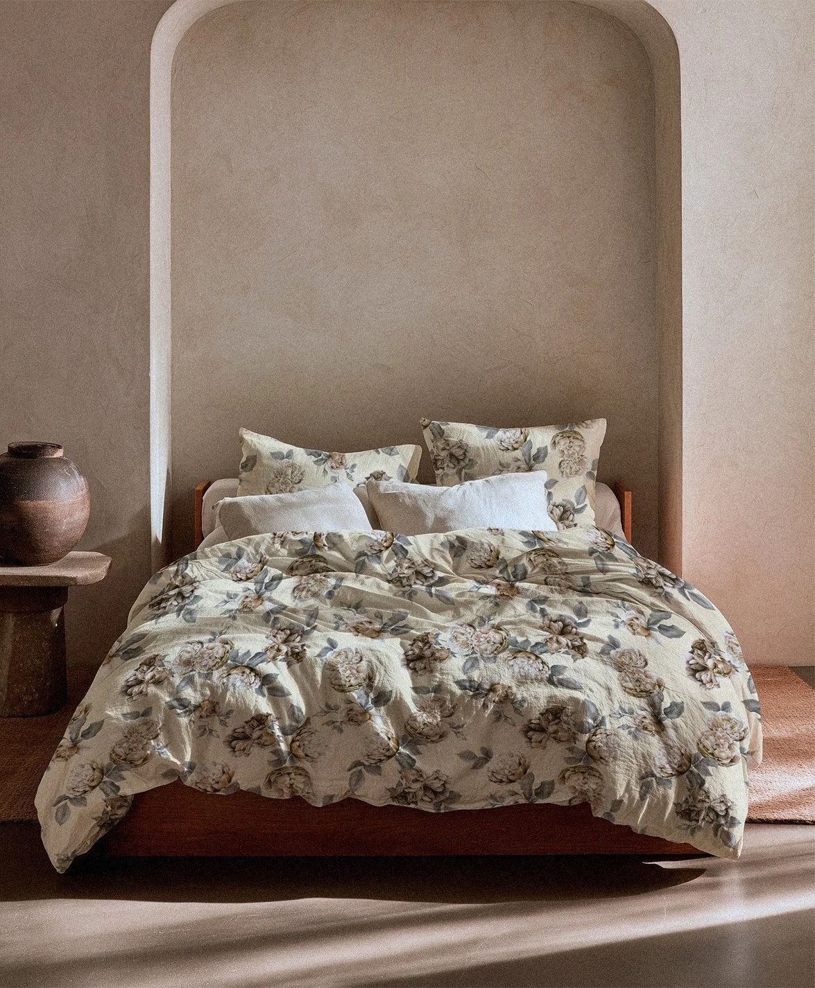 A bed with floral bedding and pillows in a minimalist bedroom with beige walls and a niche behind the bed, side table with a rustic vase.