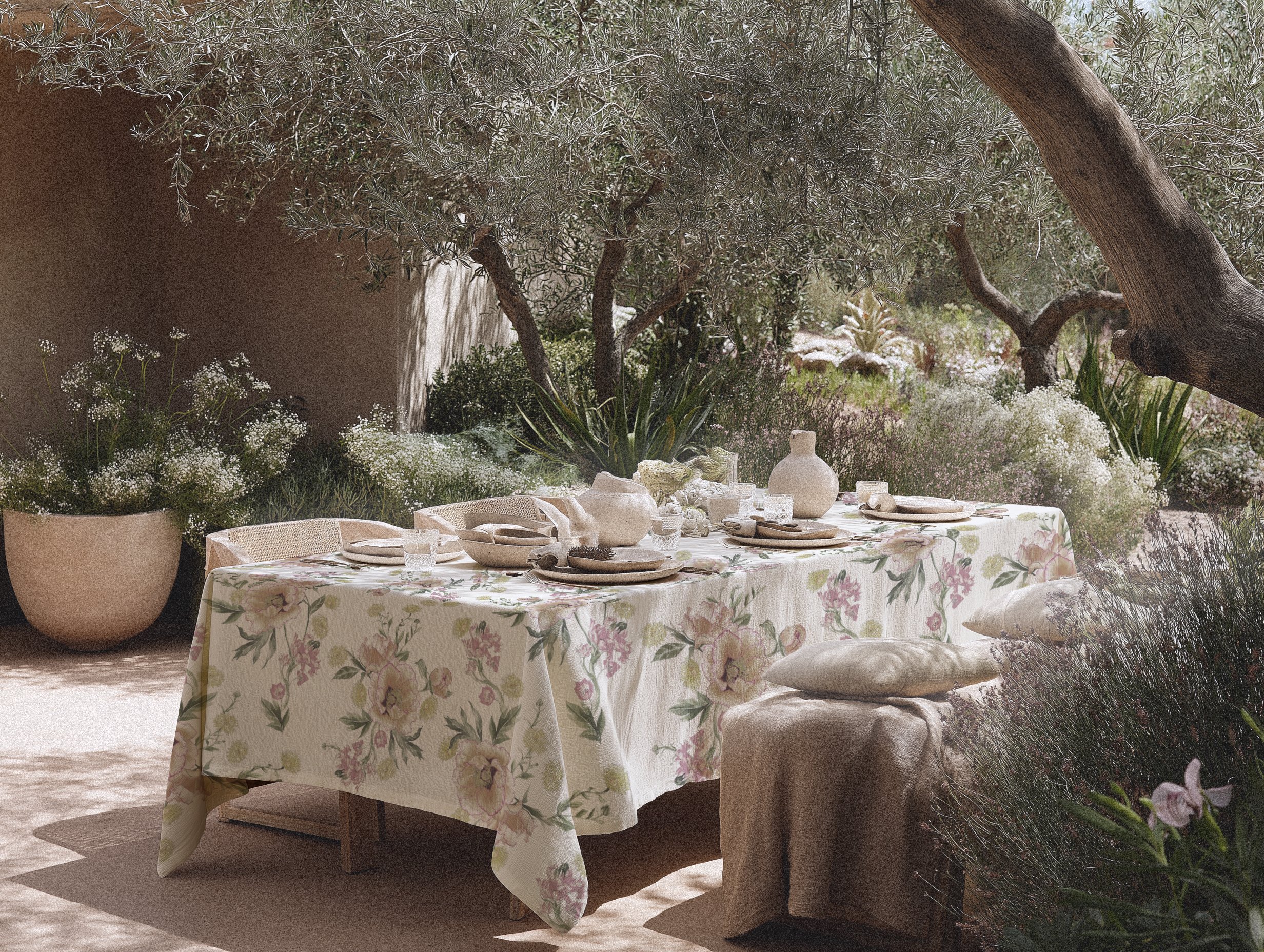 An outdoor dining table with a floral tablecloth, surrounded by plants and trees, set with plates, glasses, and ceramic vases, in a garden setting.