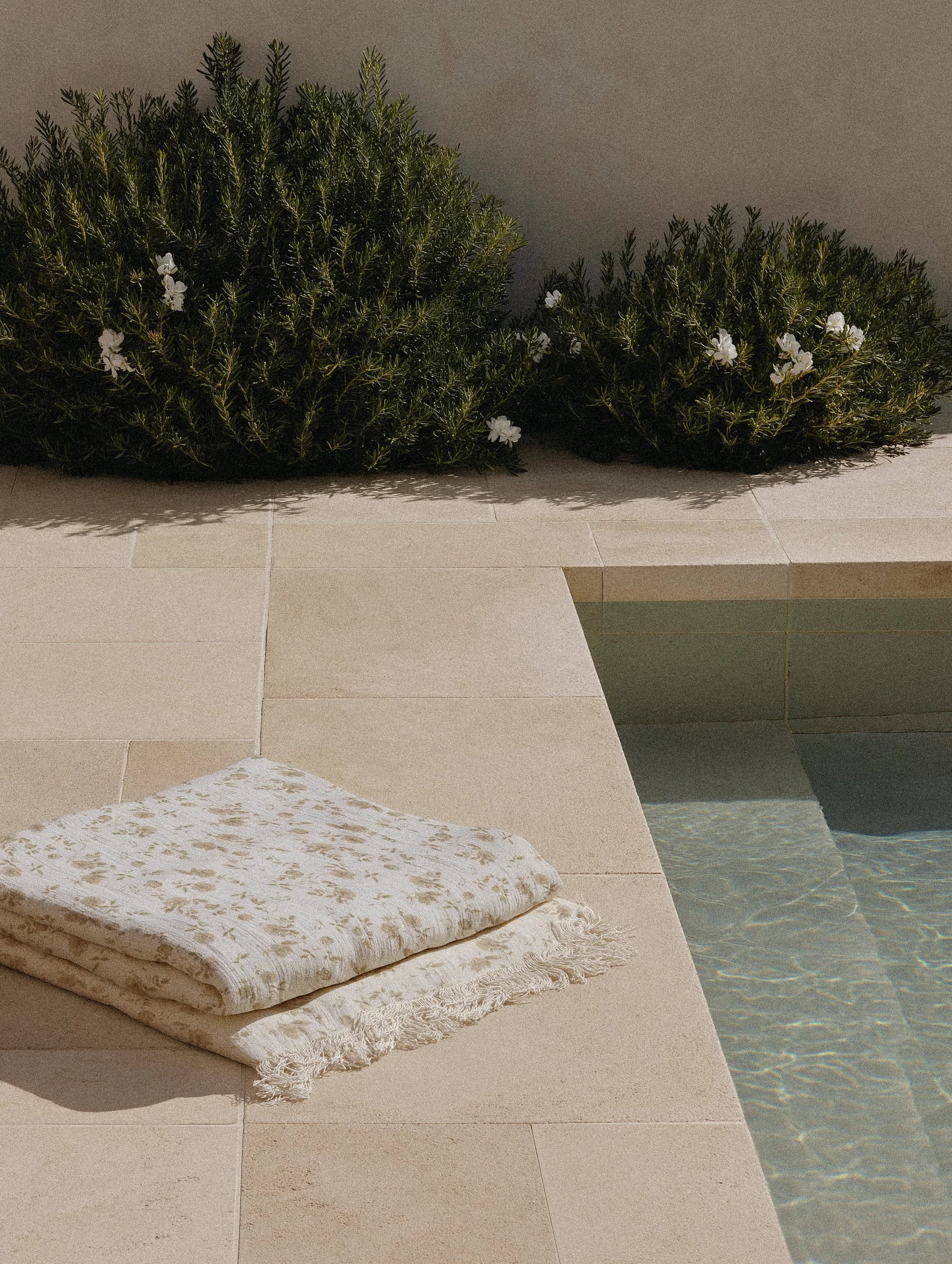 Poolside setting with beige tiles, a white towel with a floral pattern, two lush shrubbery bushes with small white flowers, and part of a swimming pool with a tiled edge and water.