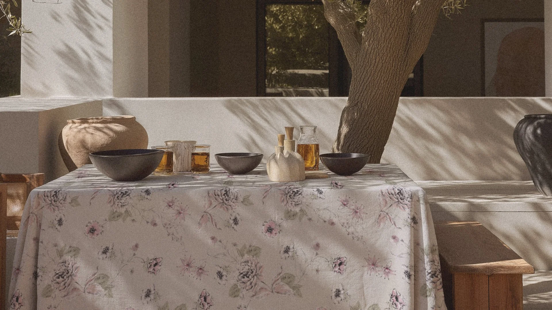 A table with a floral tablecloth, black bowls, glass bottles with amber liquid, and small vases, set outdoors with a large tree and shadows on the wall in the background.